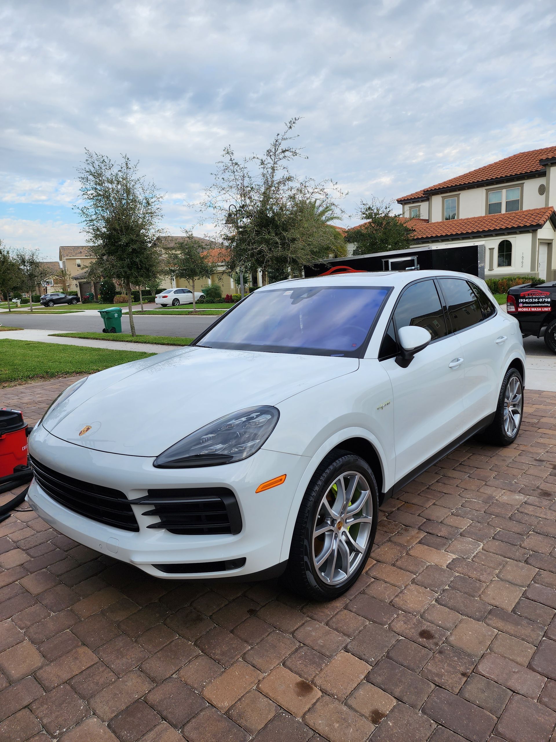 White Porsche Cayenne SUV parked on a brick driveway, with a house and cloudy sky in the background.