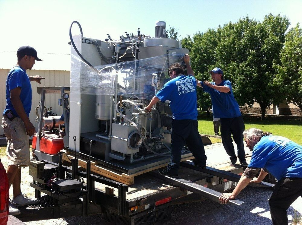 Four workers in blue shirts maneuver a large industrial machine on a trailer outdoors.