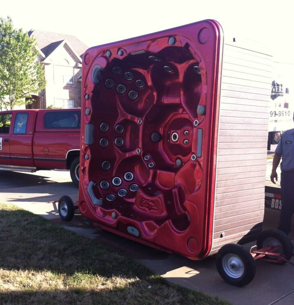 A bright red hot tub tipped on its side, secured on a wheeled dolly on a driveway with a truck in the background.