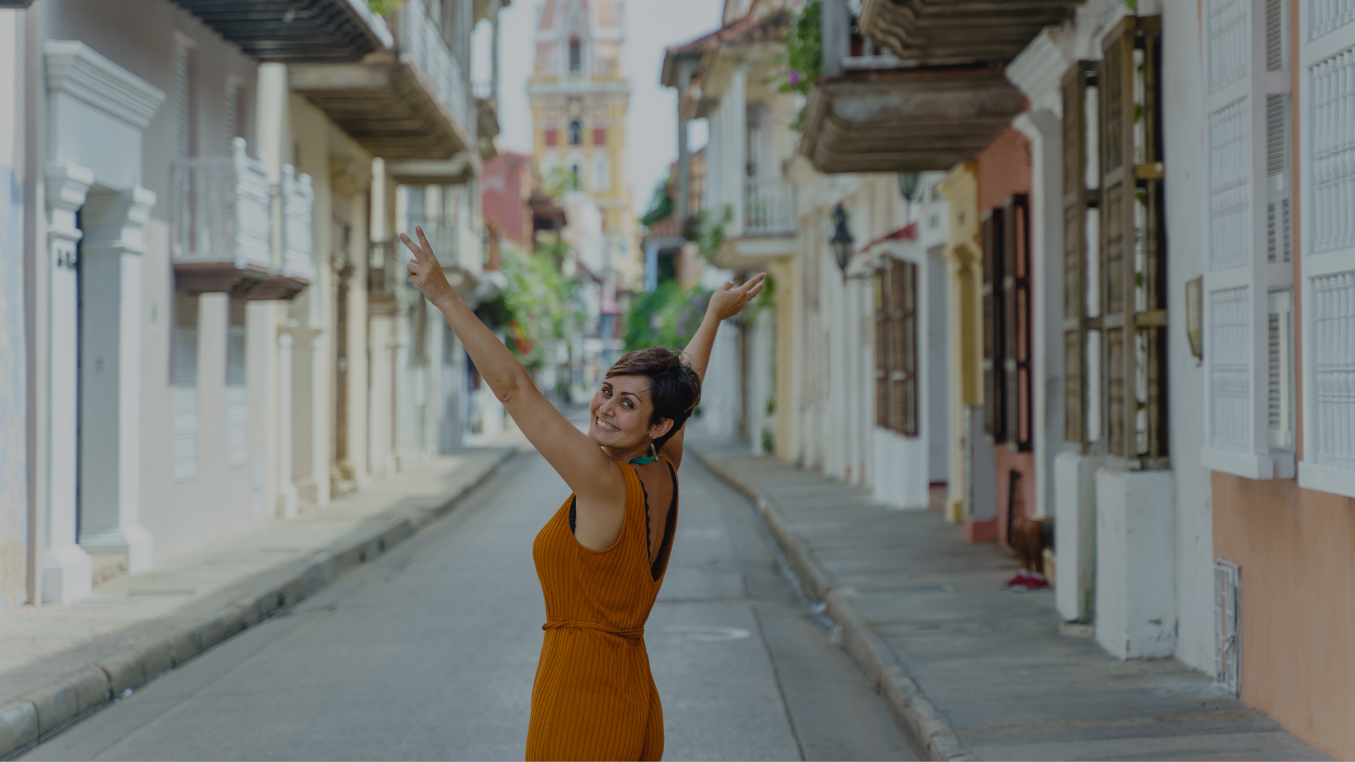 Woman in orange dress with arms raised on a colorful street, enjoying the view.