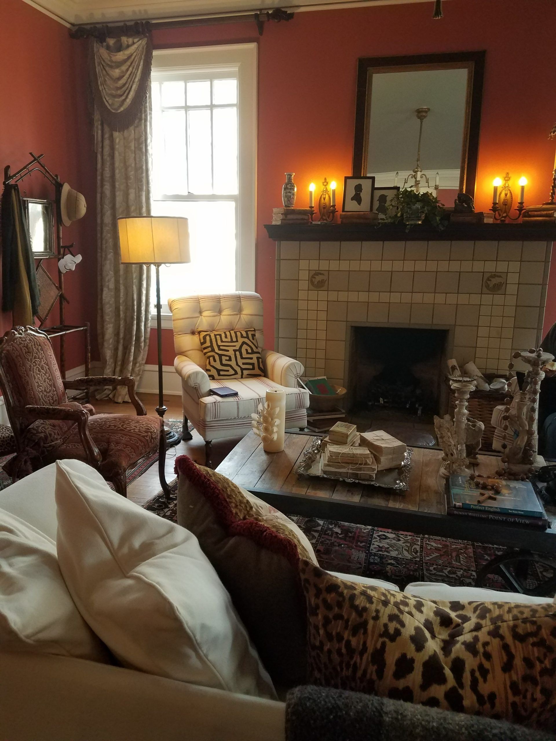 Cozy living room with fireplace, red walls, lit candles, patterned furniture, and natural light from a window.