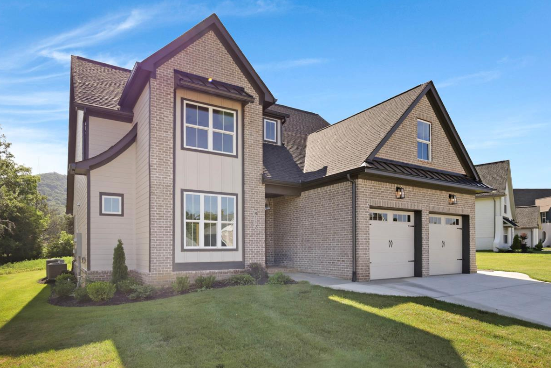 Two-story house with tan siding, black trim, two-car garage, and green lawn under a blue sky.