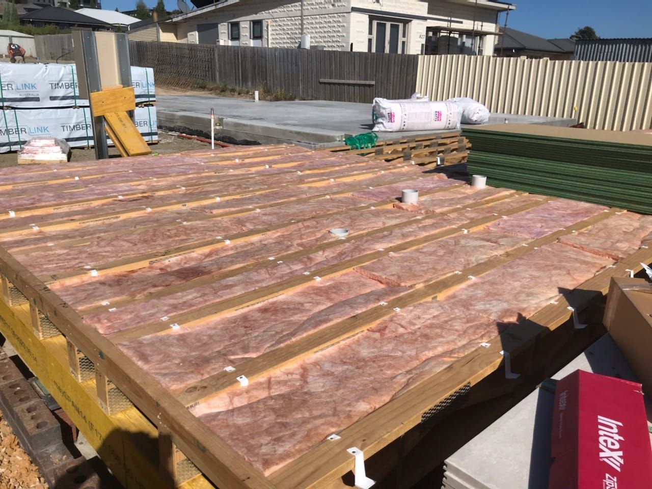 Wooden decking panels stacked on a trailer at a construction site, with materials and a fence in the background