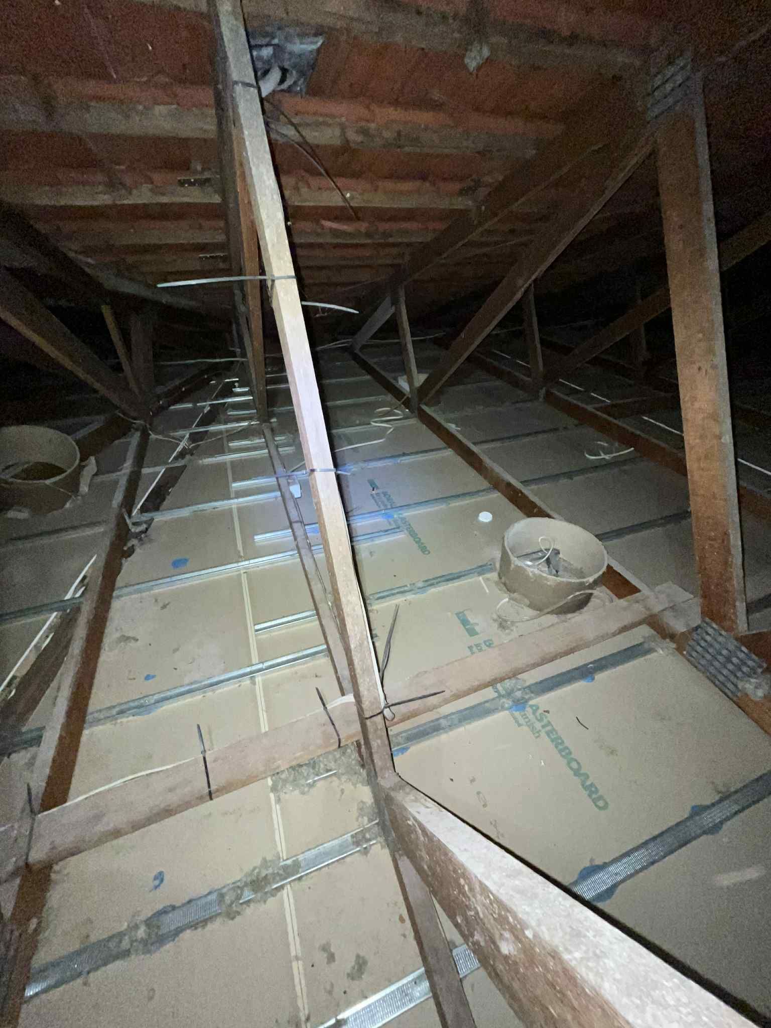 Dim attic with exposed rafters, insulation batts, and a bucket on the floor.