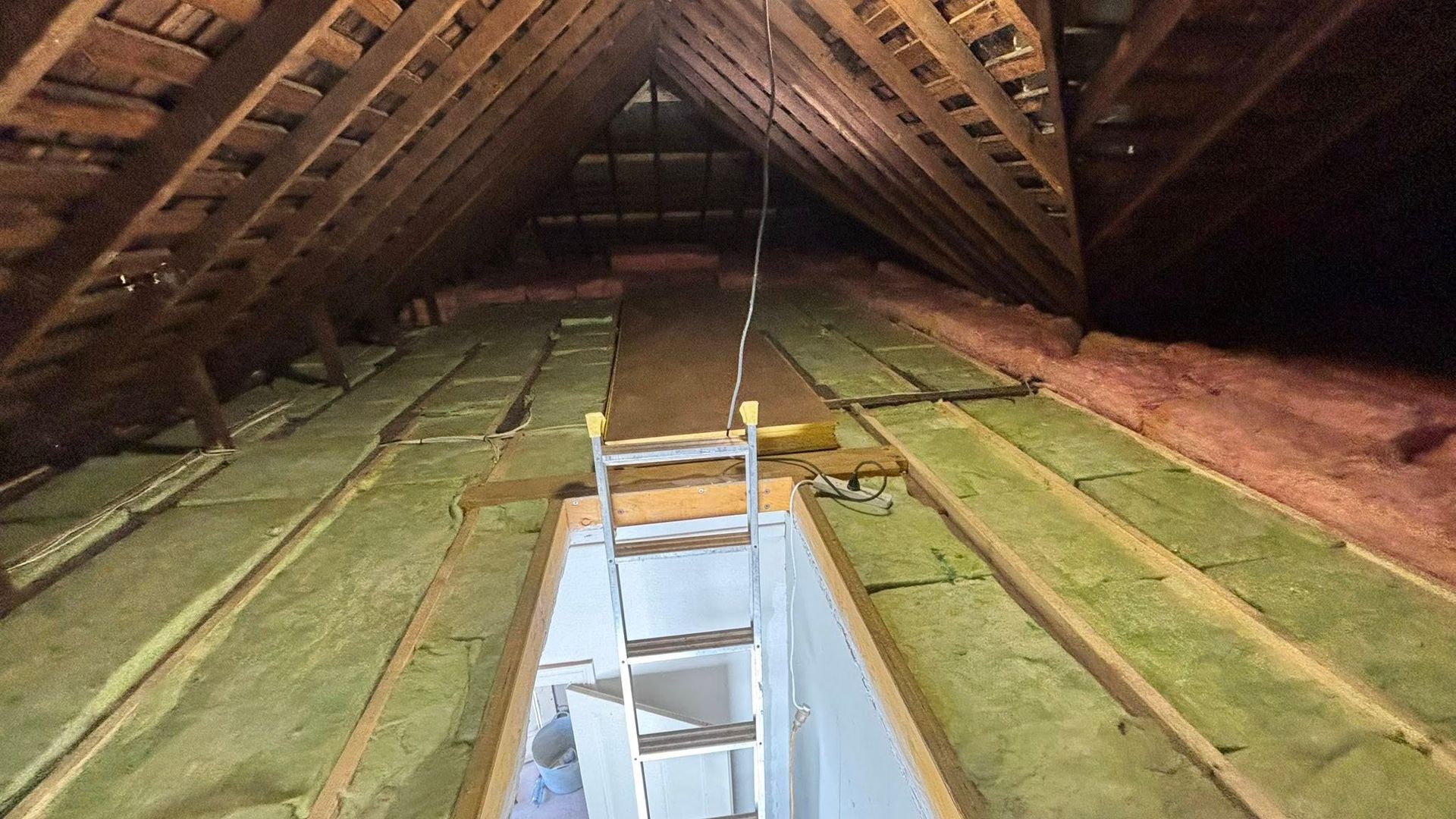 Attic interior with exposed rafters, green insulation, and an open ceiling hatch with a ladder below