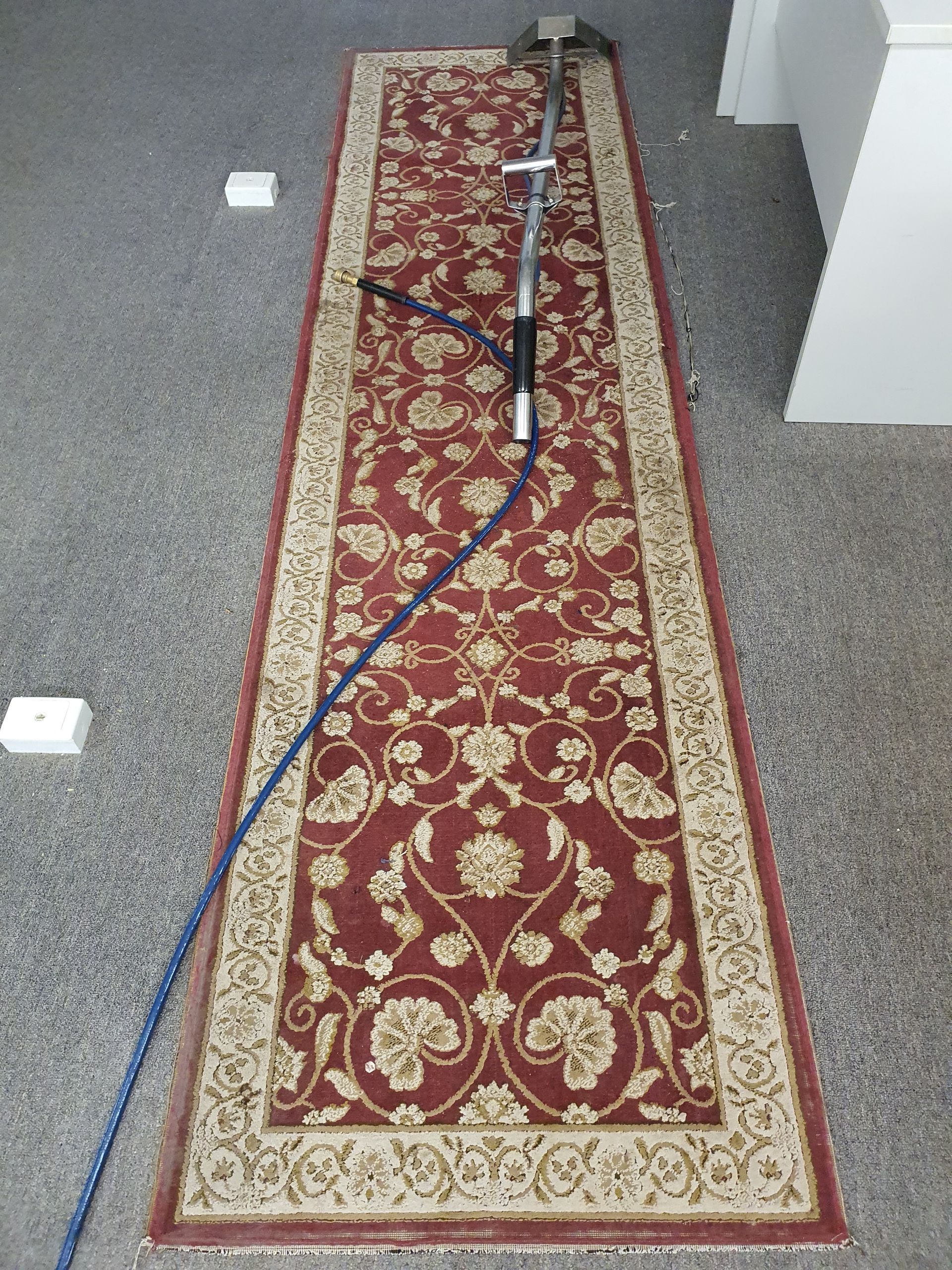 Long, red patterned rug with cleaning tool on a gray floor.
