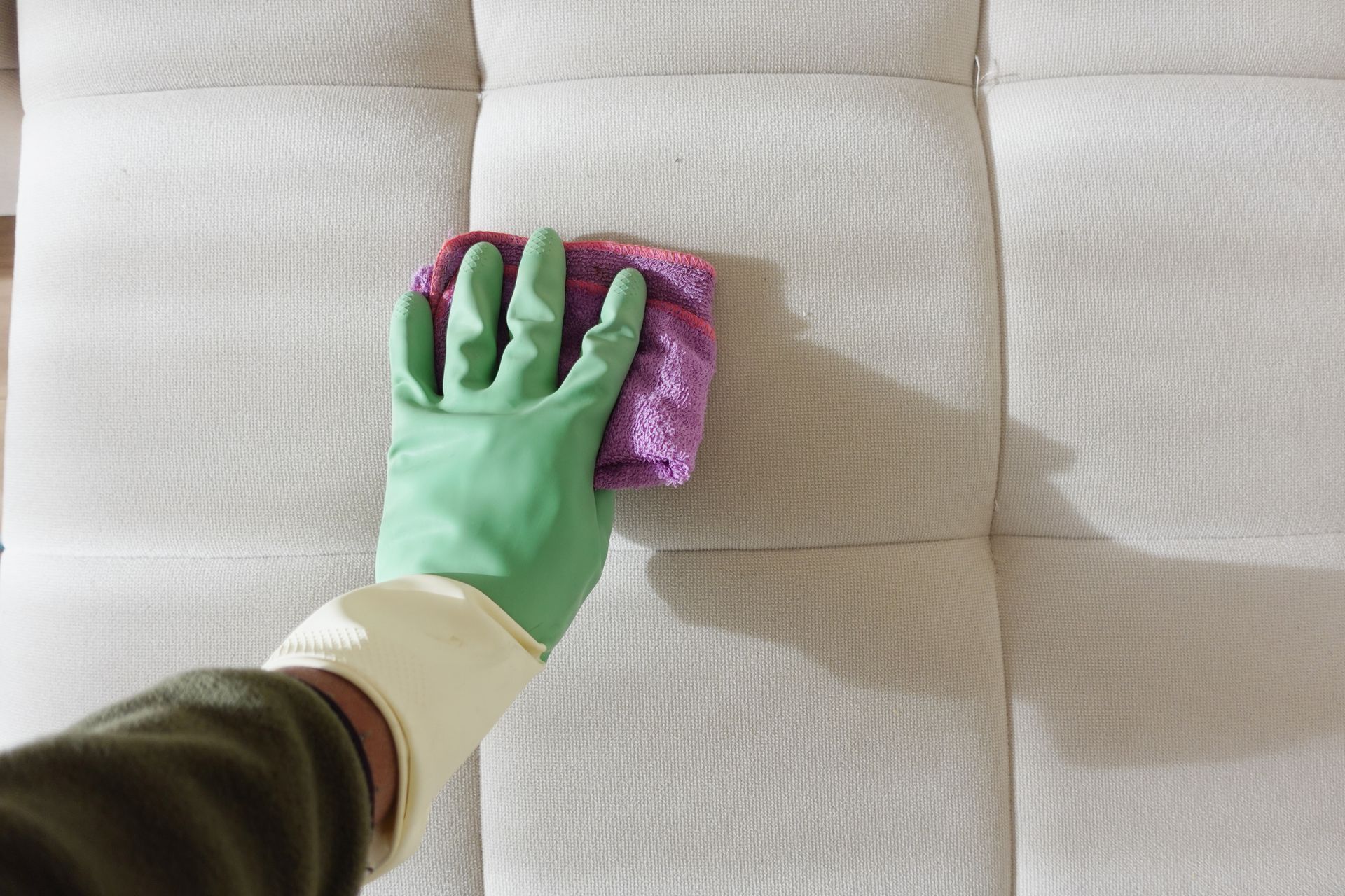 A hand wearing a green glove wipes a sofa with a purple cloth gently.