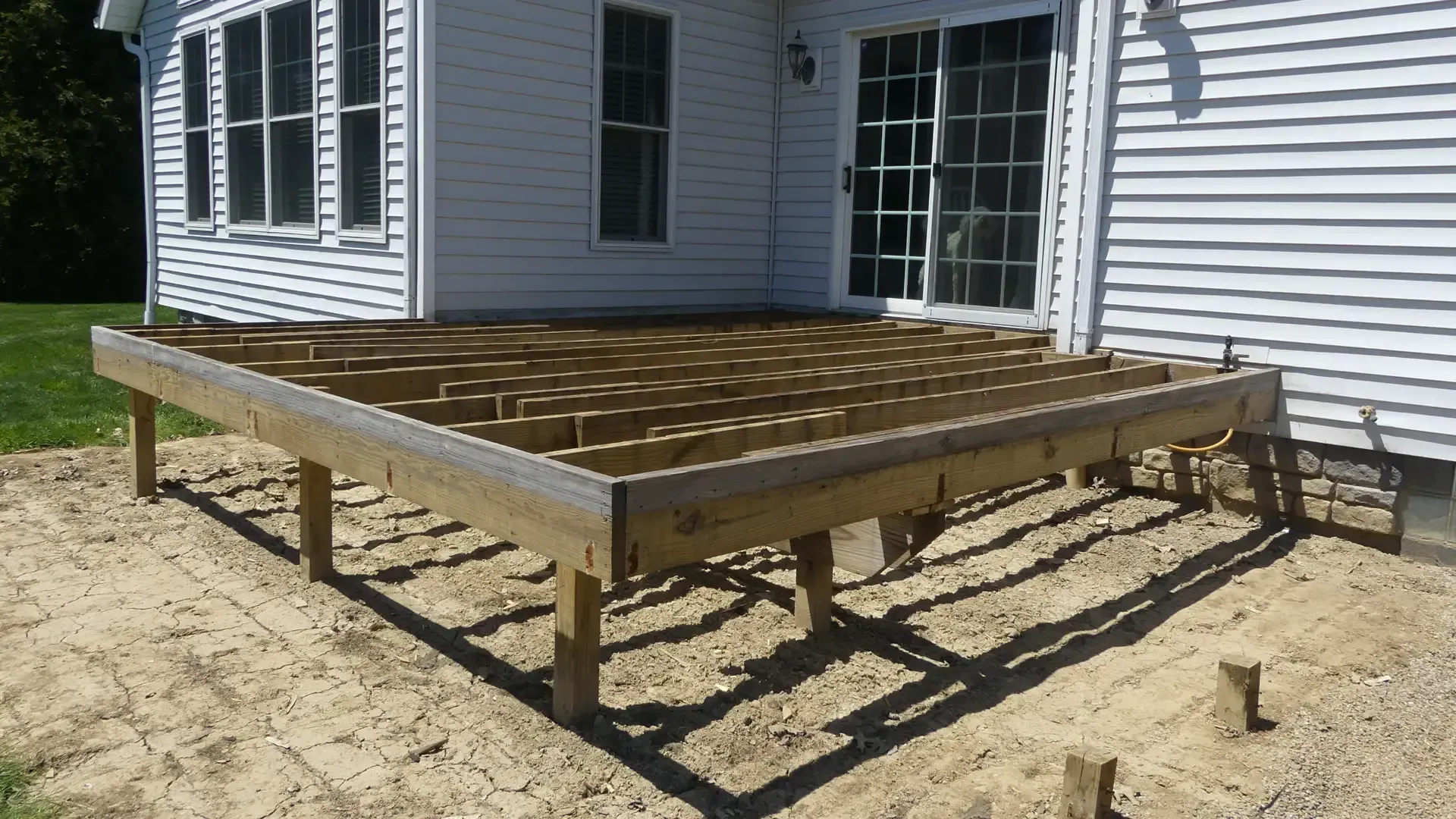 A wooden deck frame under construction attached to the side of a white house on a dirt lot.