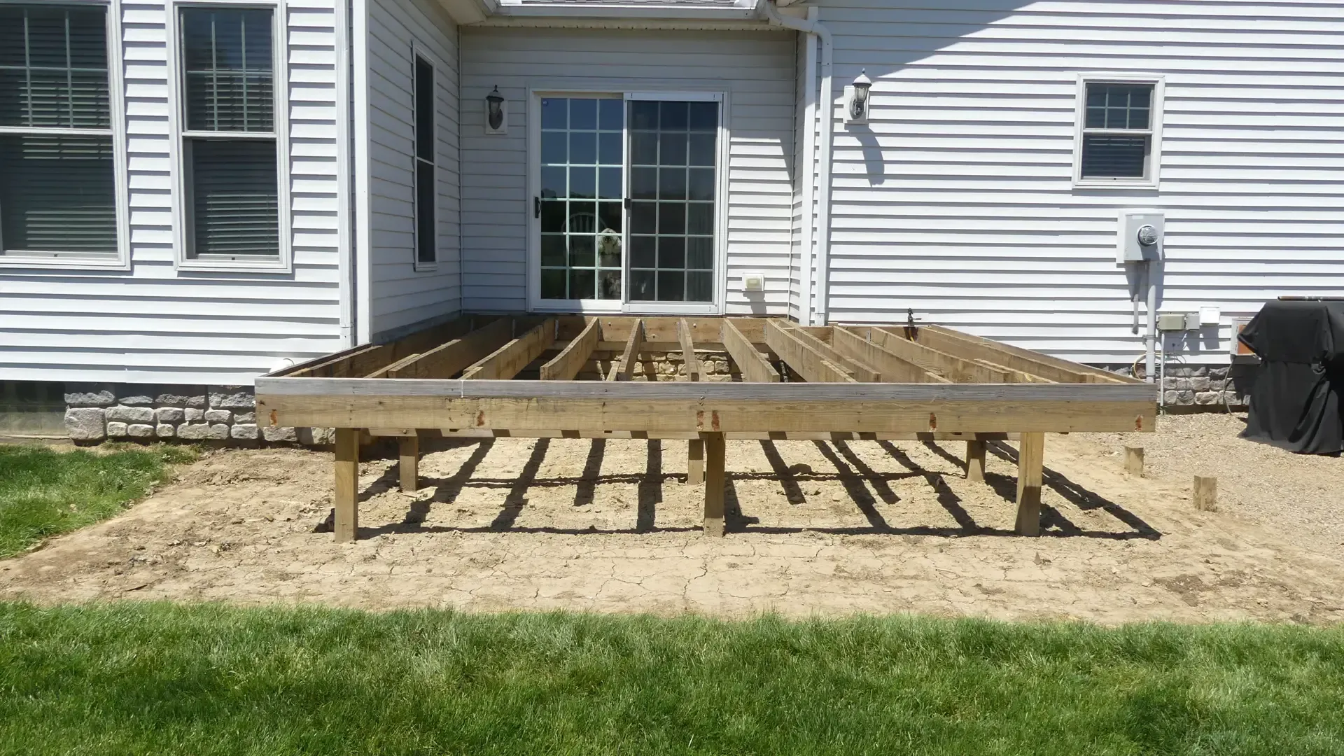 A wooden deck frame under construction attached to the back of a house with white siding over a gravel ground.