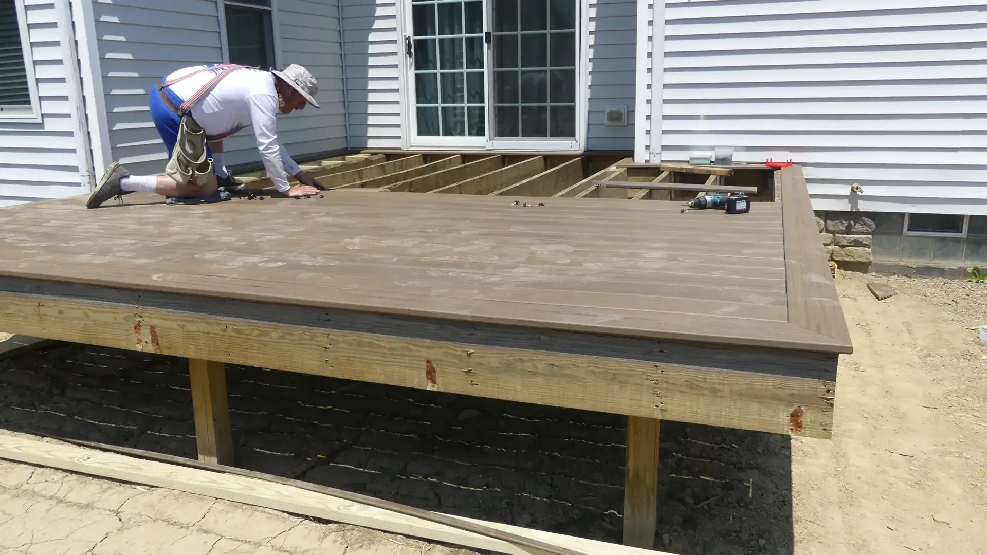 A worker in a hat and safety harness installs composite decking boards on a residential backyard deck frame.