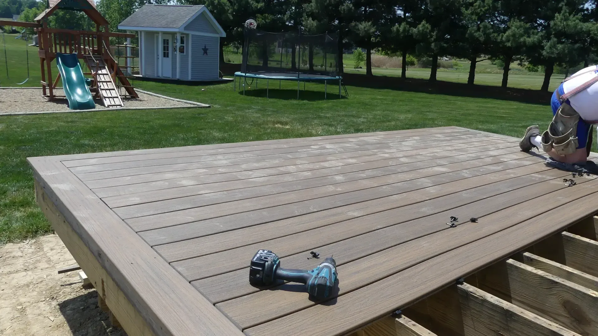A partially constructed wooden deck in a grassy backyard with a drill sitting on the surface.