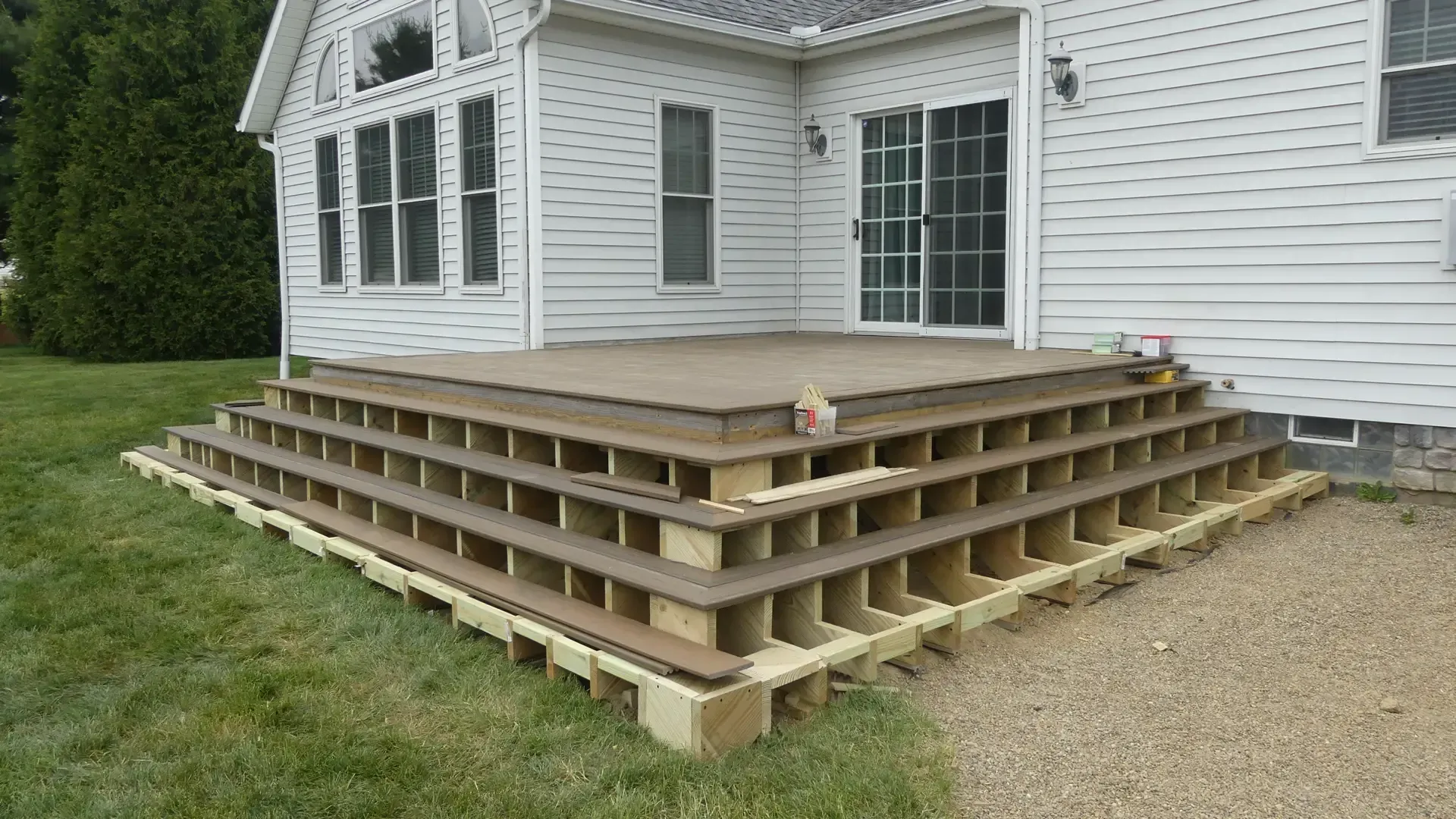 A wooden deck under construction outside a white-sided house, showing exposed support framing beneath tiered steps.