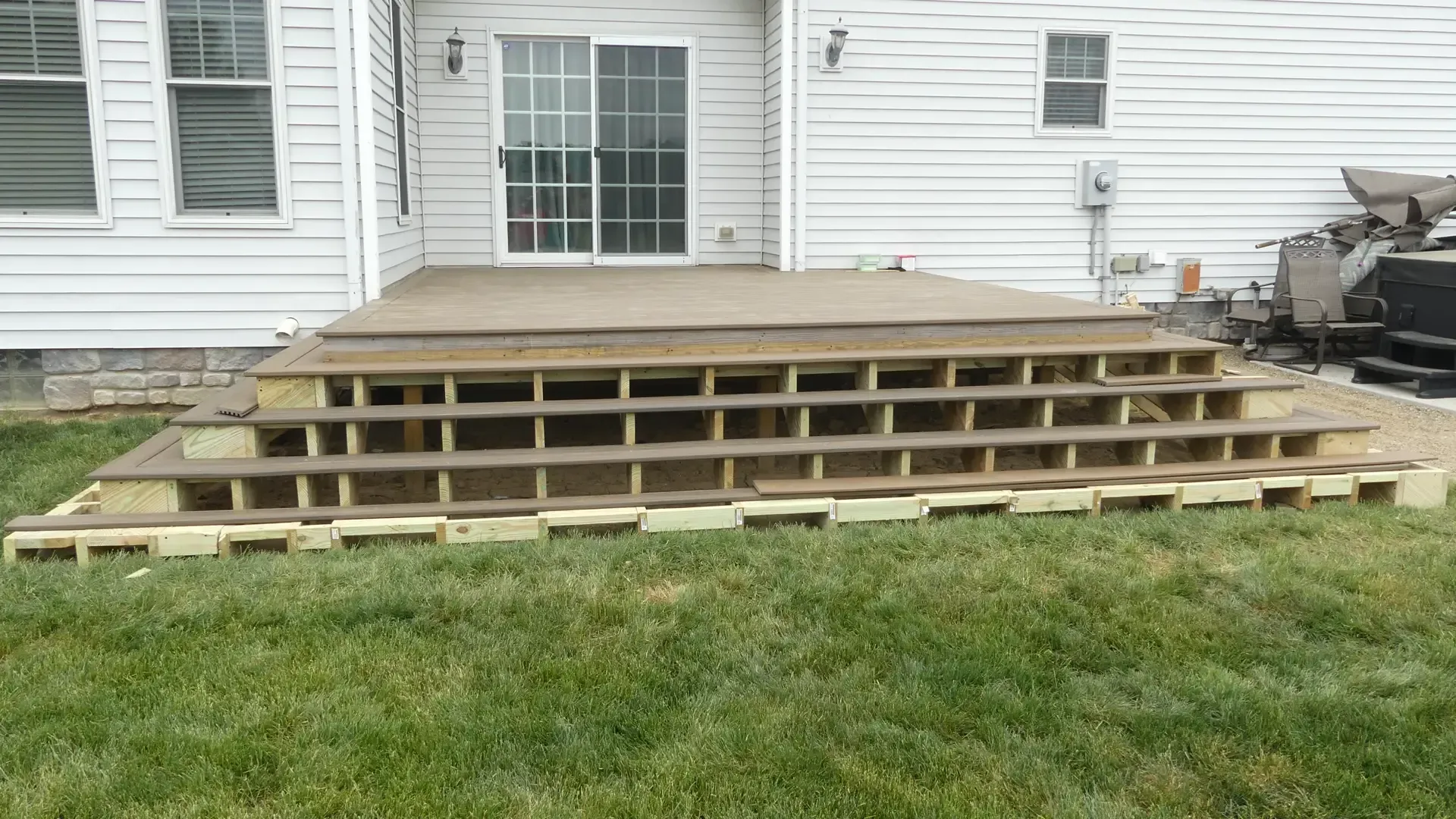 A partially finished wooden deck with open-riser steps sits against the white siding of a house in a grassy backyard.
