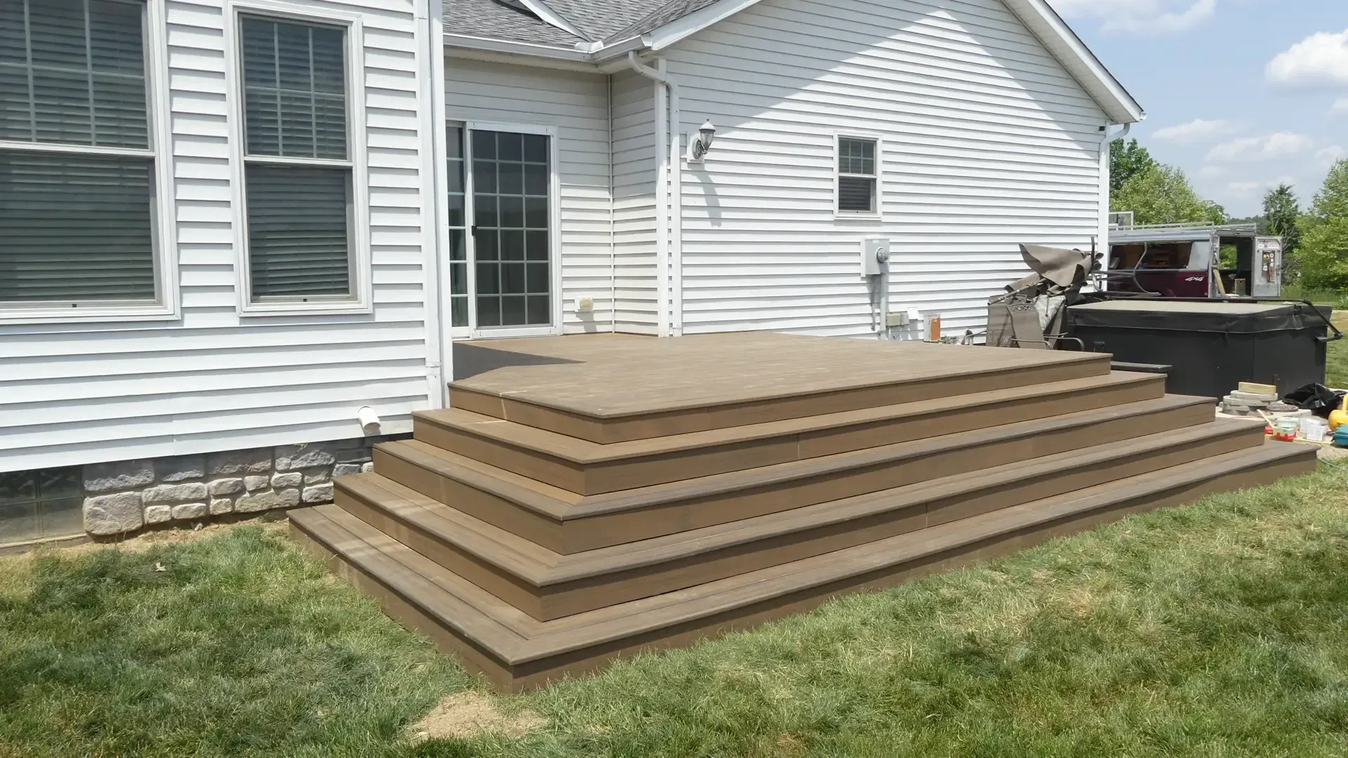 A wide, multi-level brown composite deck extends from the back of a white-sided house with a grass lawn.