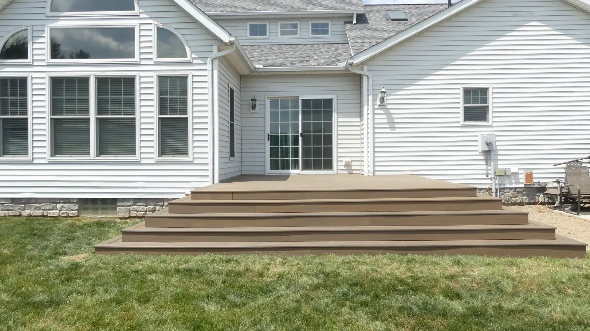 A wide, brown deck with several steps leading up to the sliding glass back door of a white-sided house with a grassy lawn.
