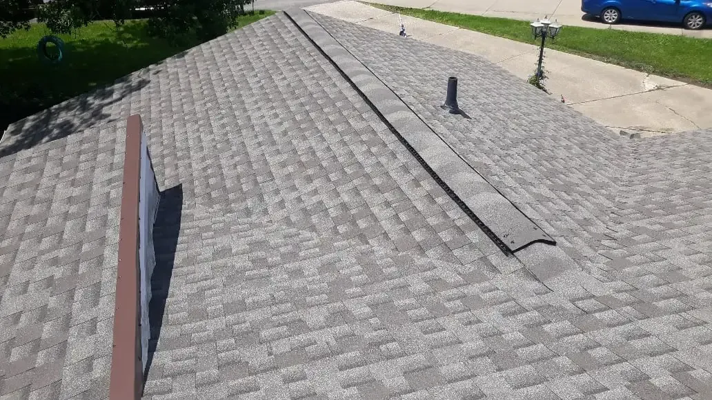 A view of a grey shingled roof with a ridge vent and a plumbing pipe, taken from an elevated perspective.