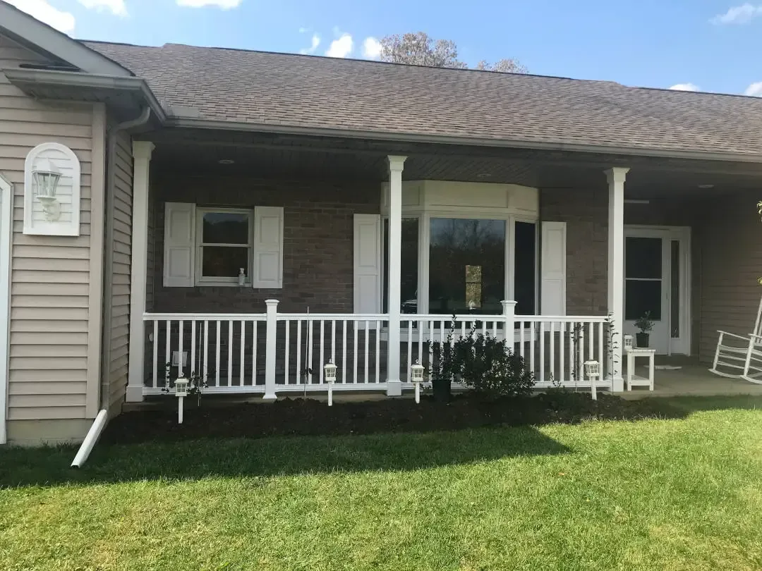 A one-story suburban home with brick siding, a white porch railing, and a front lawn under a bright blue sky.
