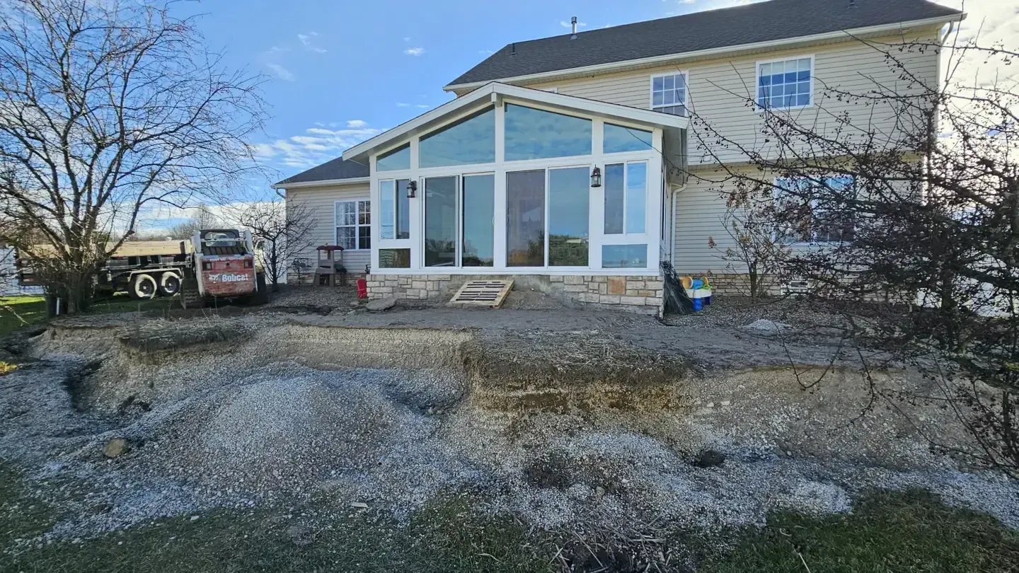 A two-story tan house with a large new sunroom addition and a construction site with gravel in the yard.
