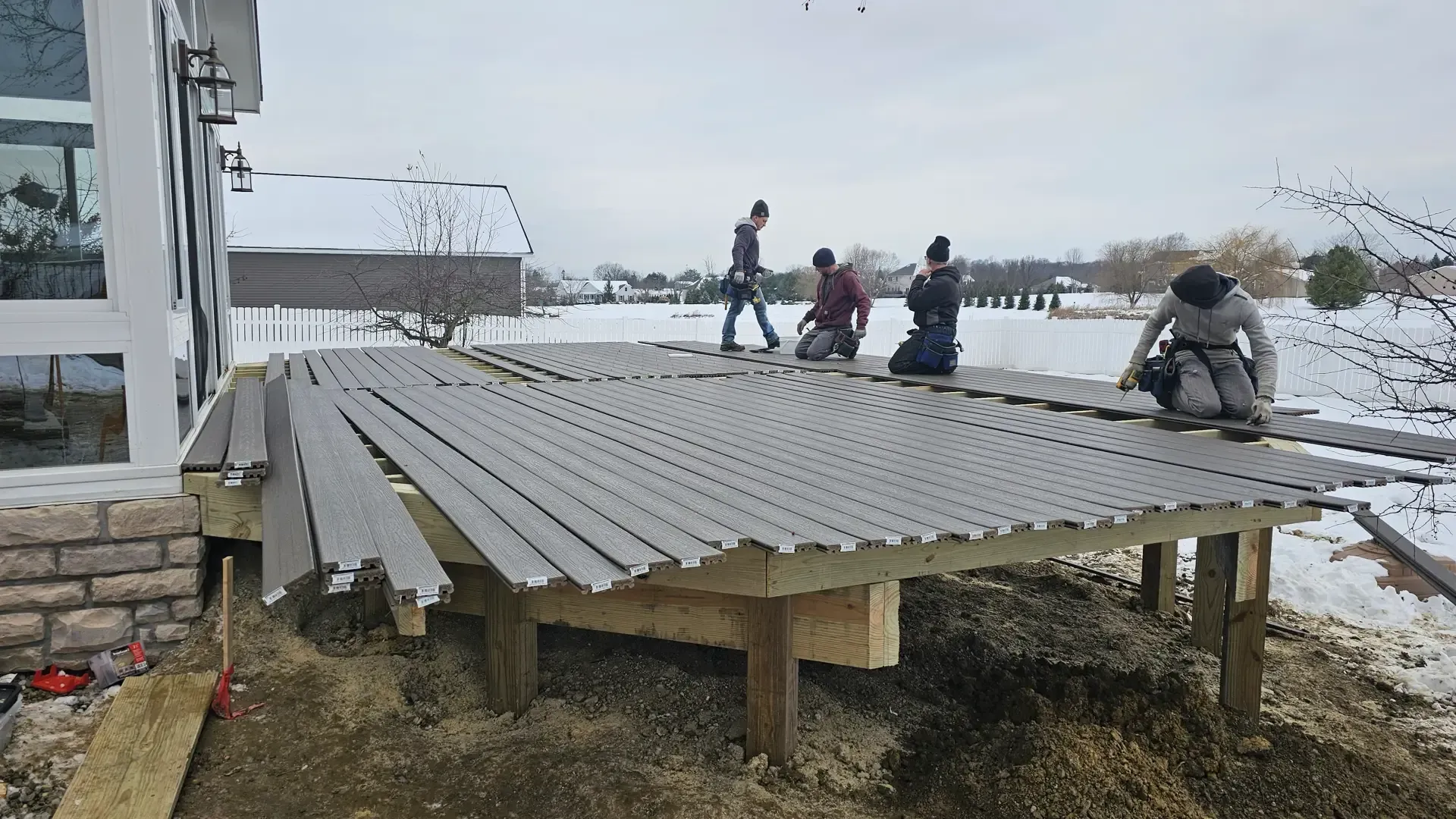 Four people work on a curved composite deck installation outside a house during winter.
