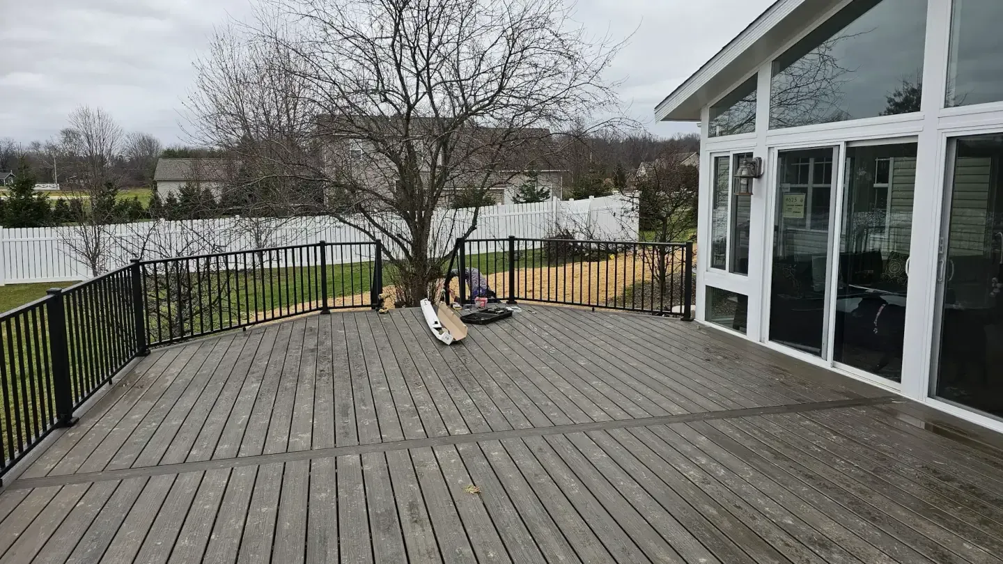 A wooden deck with a black metal railing outside a house with large windows, overlooking a backyard with a white fence.