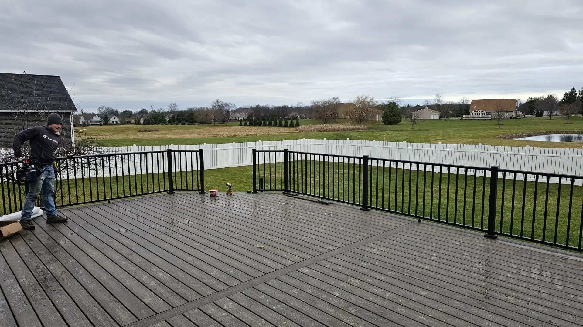A person installs black metal railing on a gray wood deck overlooking a grassy field with a white fence.