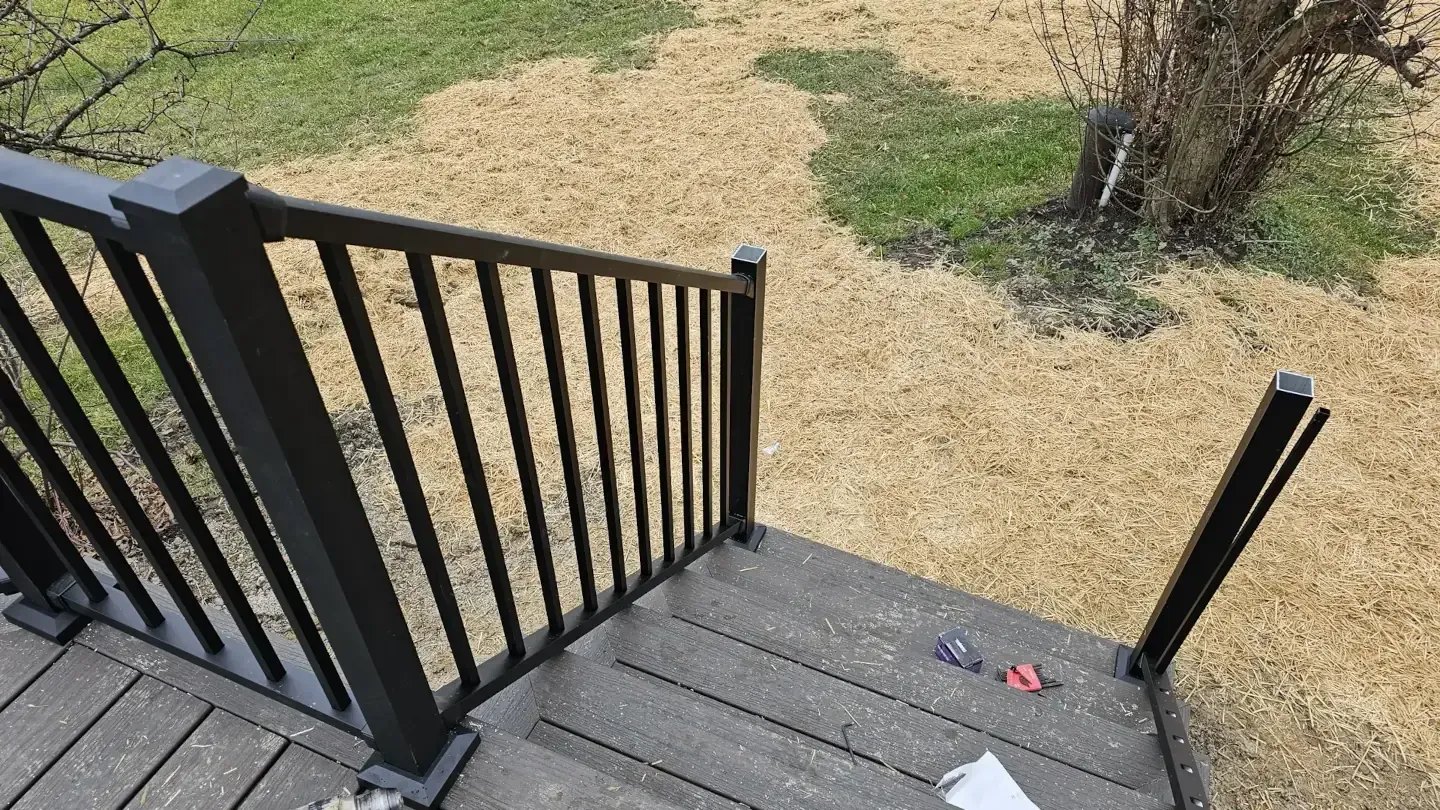 Black aluminum deck railing with vertical balusters leads down steps to a mulch-covered yard.