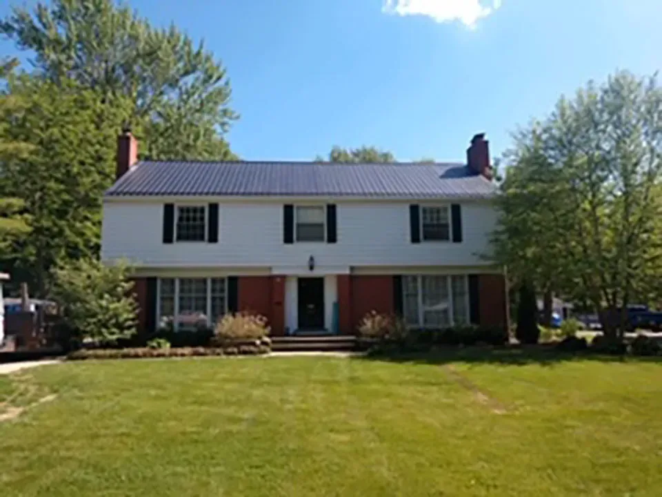 A two-story house with white siding on the top, a red brick bottom, and a dark standing-seam metal roof on a sunny day.