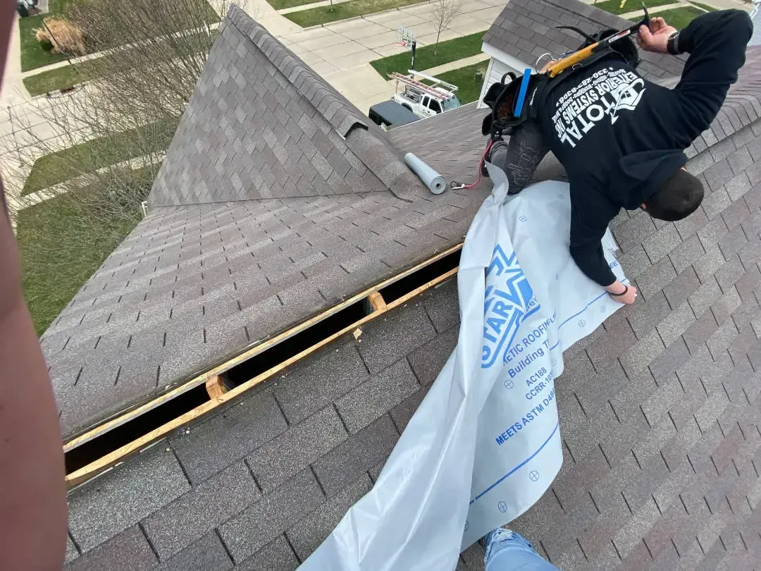 A person in a black hoodie installing roof underlayment on a shingled roof with an open ridge vent.