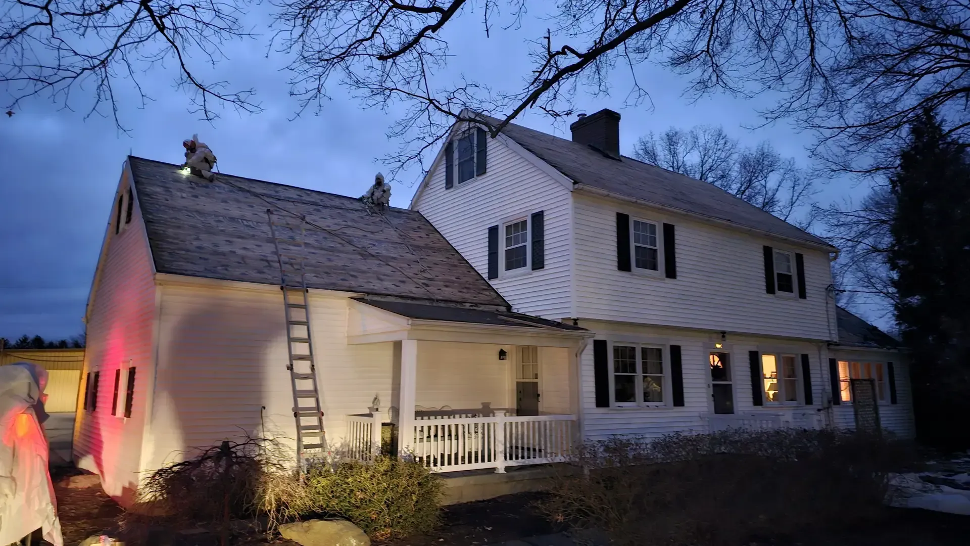 A white two-story house at dusk, featuring a ladder against the roof and festive Halloween decorations.