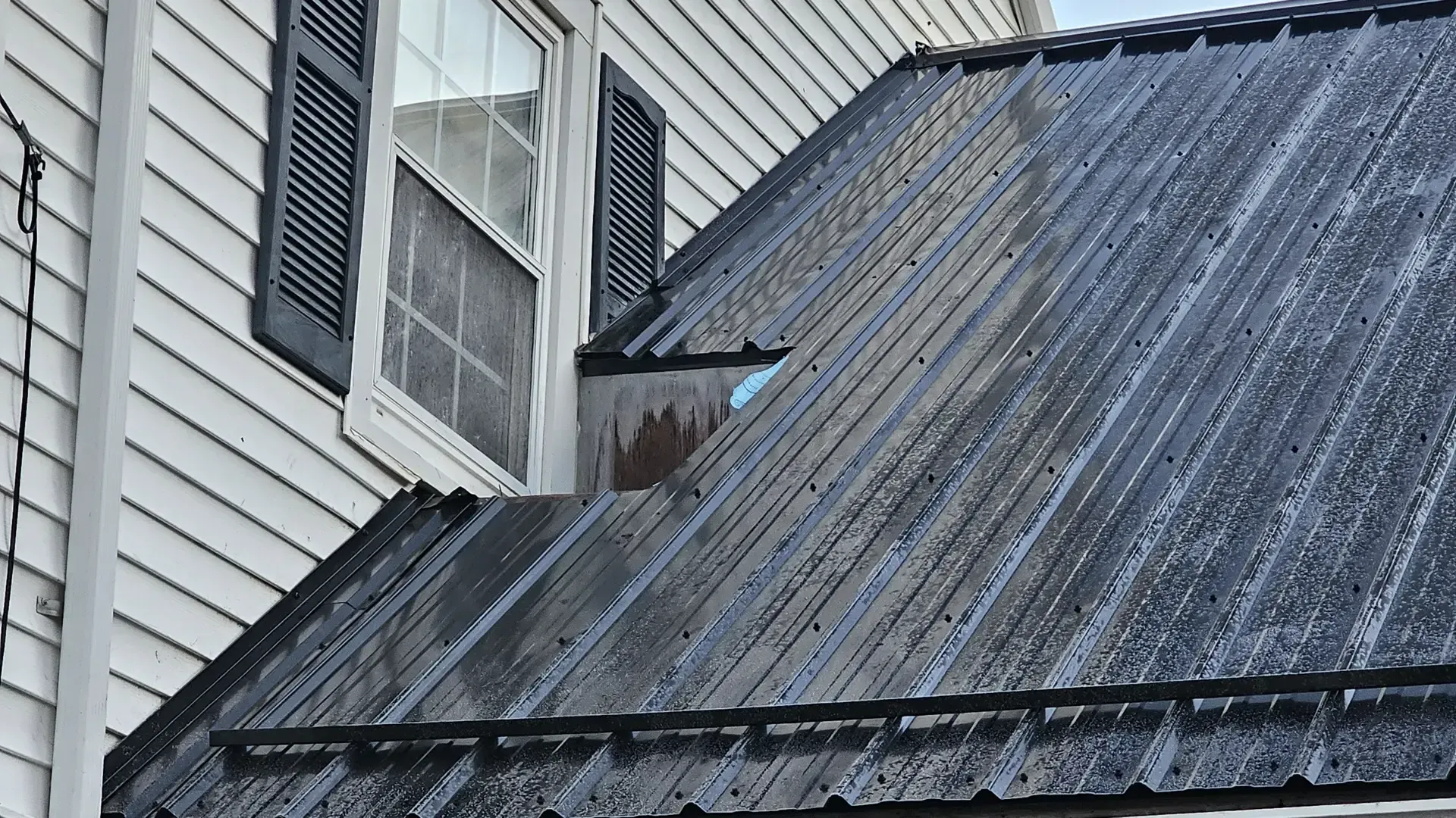 A metal roof meets the side of a house with white siding and a window with black shutters.