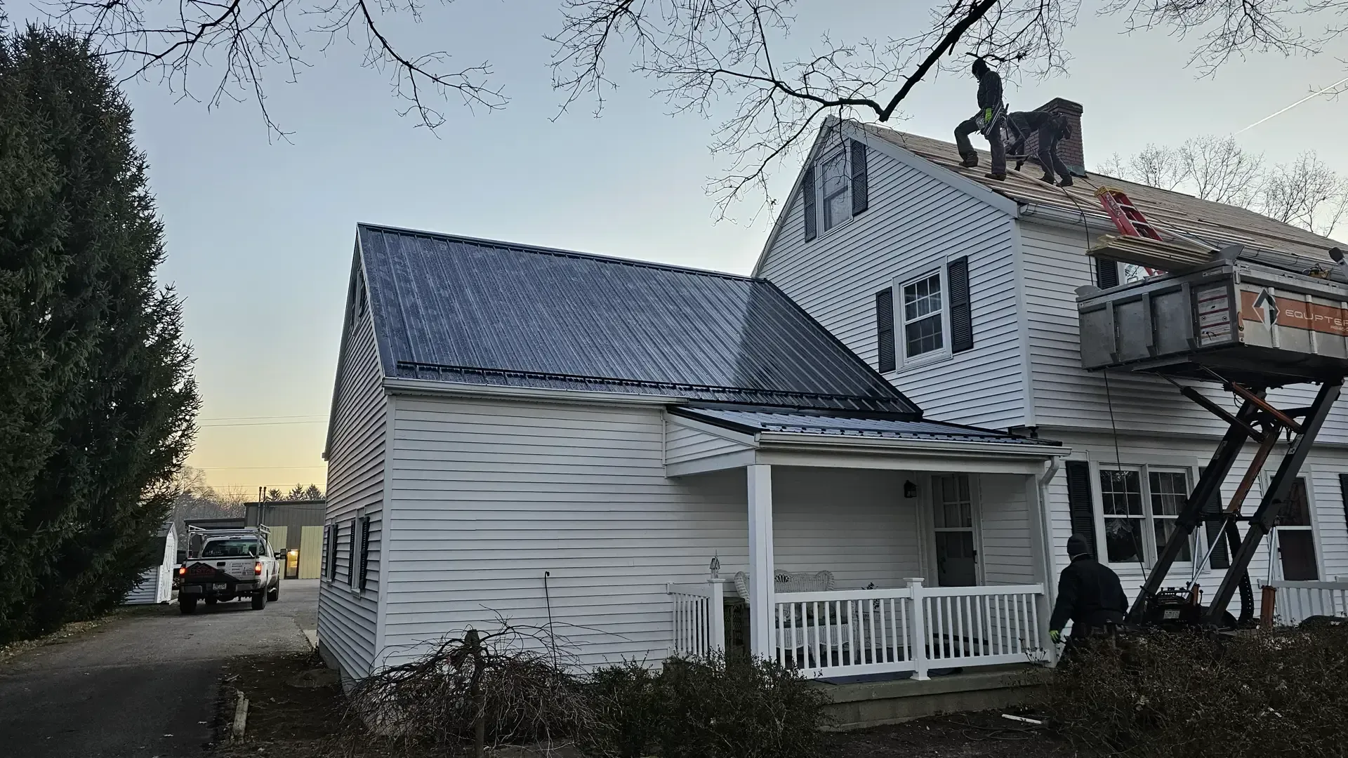 Workers installing a new dark metal roof on a white two-story house with a wooden scaffold on the side.