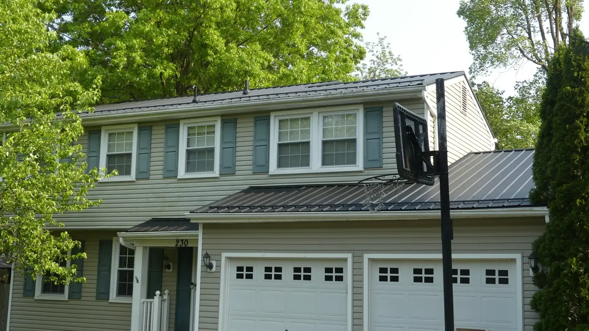 A two-story suburban house with light gray siding, dark metal roofing, and a basketball hoop in the front yard.