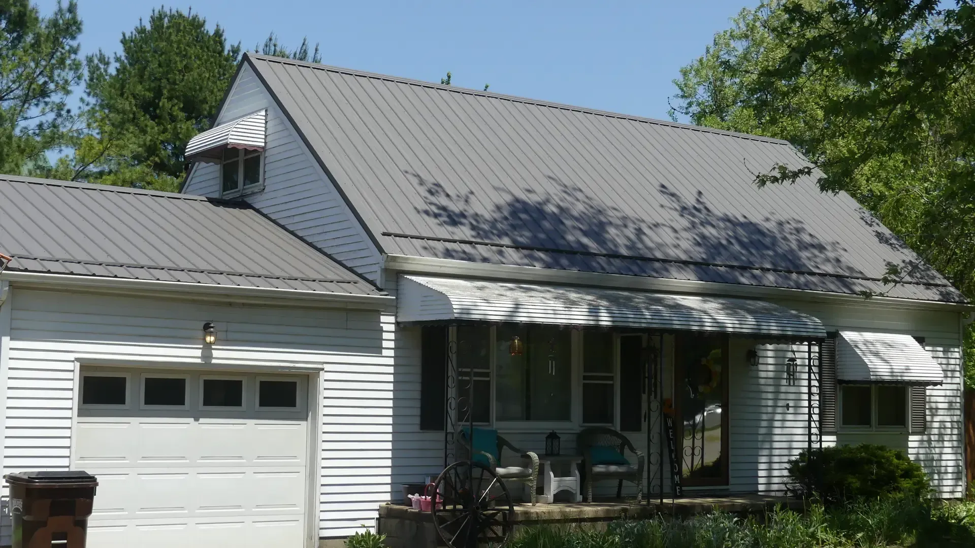 A white one-story house with a grey metal roof, an attached garage, and white window awnings surrounded by green trees.