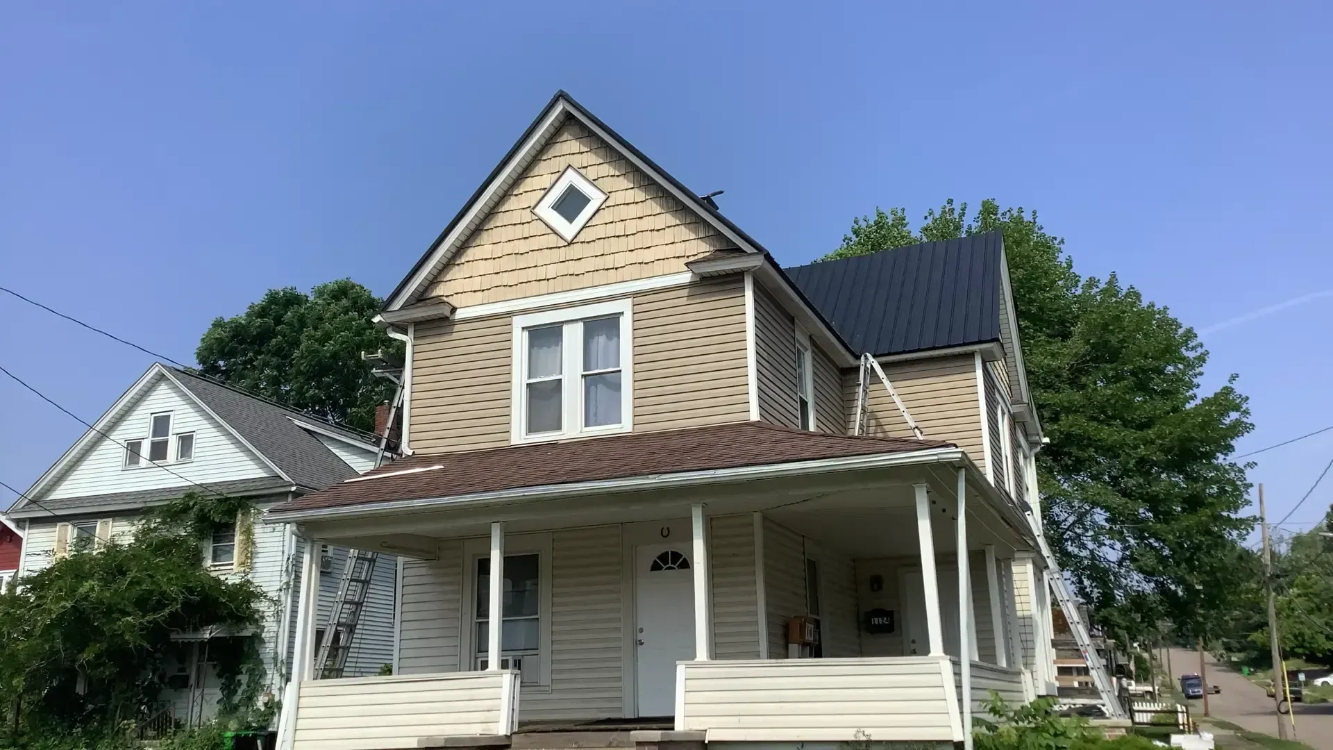 A two-story tan house with a wrap-around porch and a dark metal roof under a clear blue sky.
