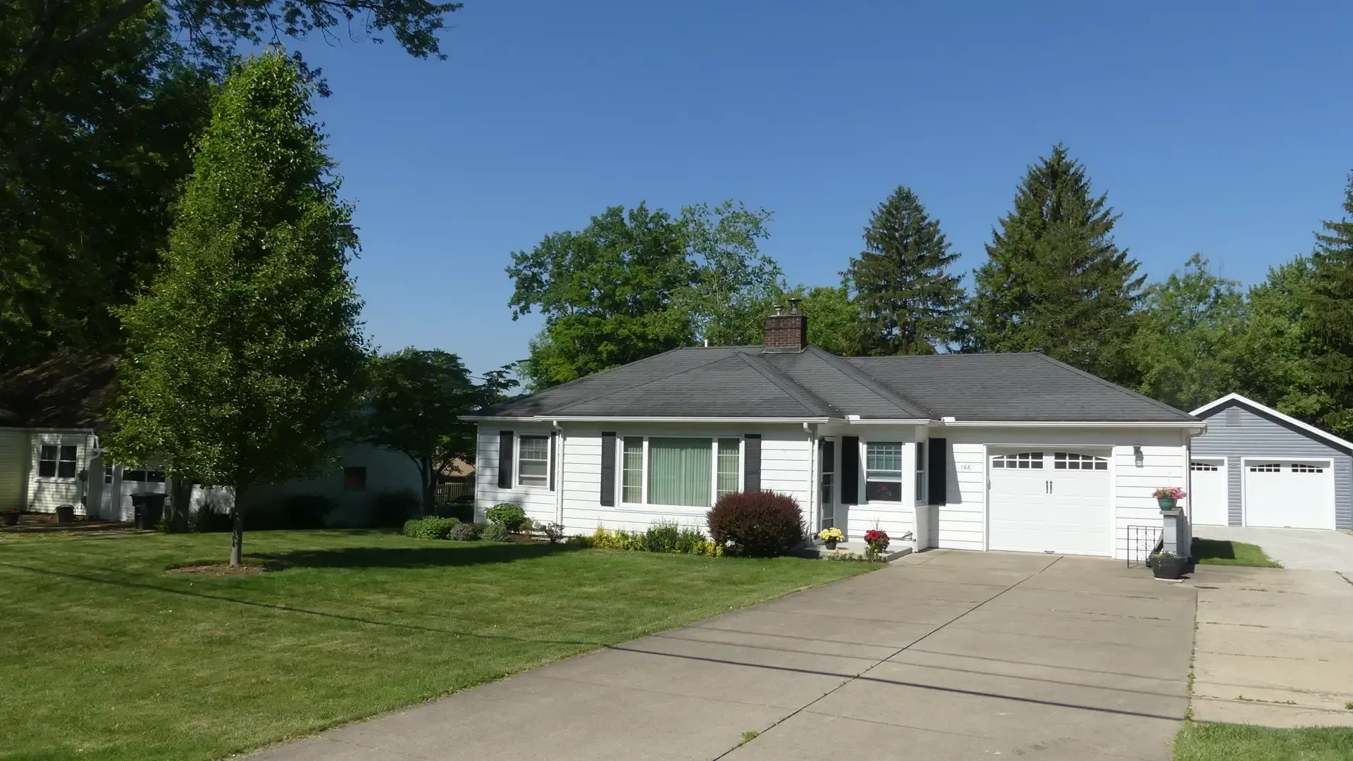 A white, single-story ranch-style house with a gray roof, large lawn, and detached garage on a sunny day.