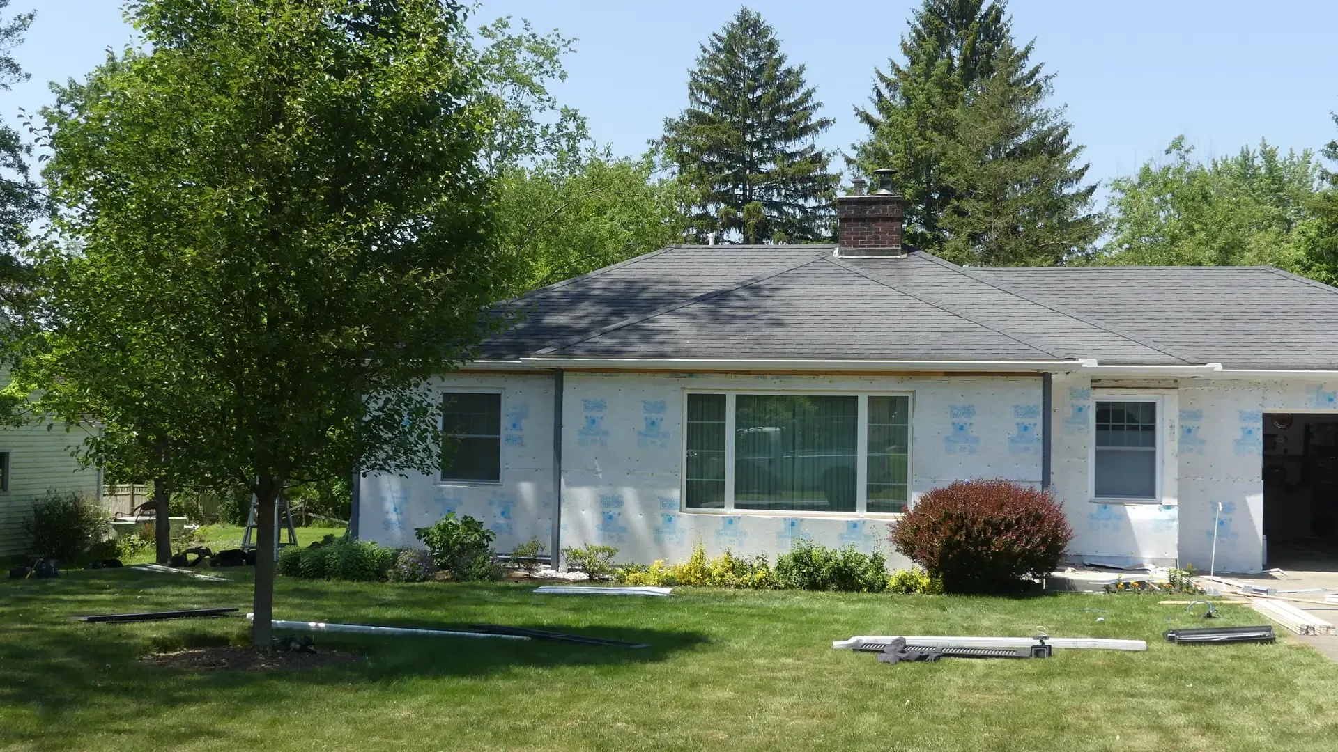 A single-story house undergoing exterior renovations, featuring white insulation boards, a shingled roof, and a front lawn.