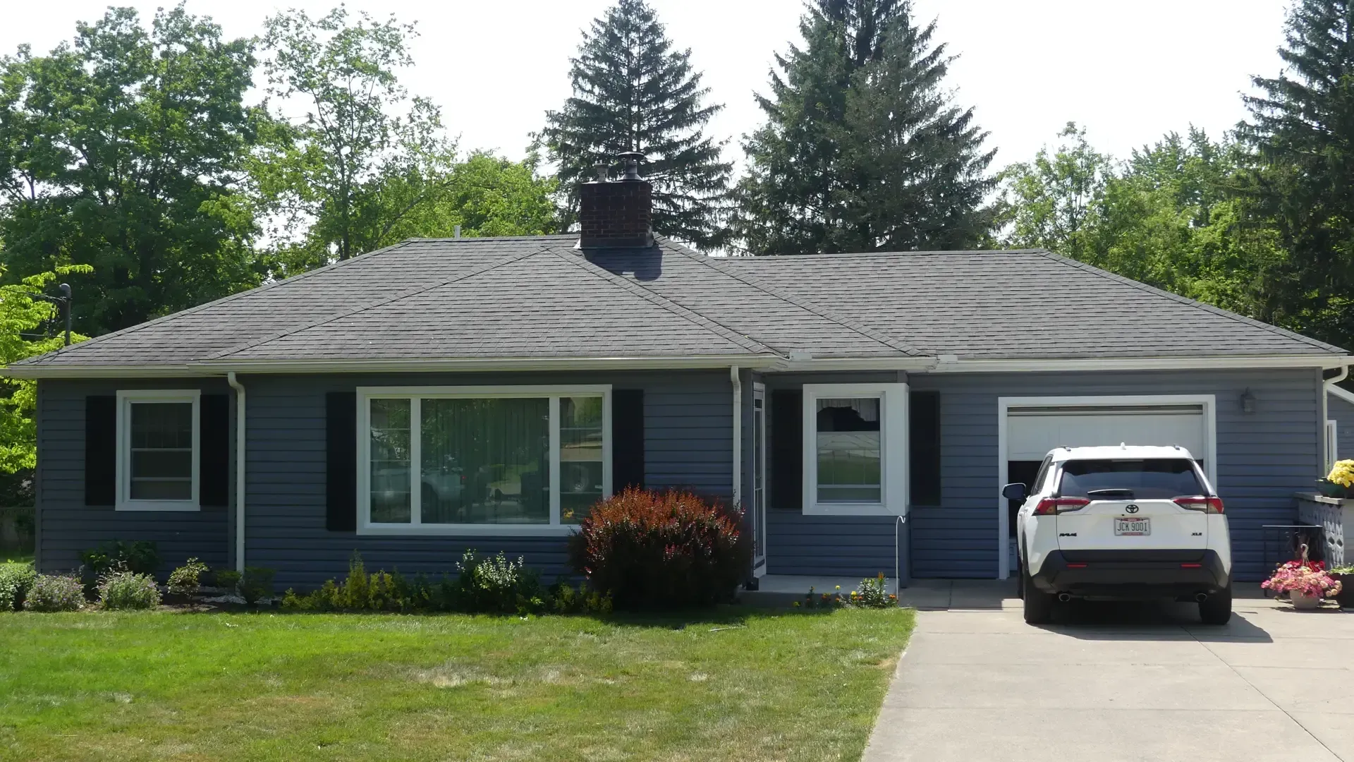 A blue, single-story suburban house with a gray shingled roof, a front lawn, and a white SUV parked in the attached garage.
