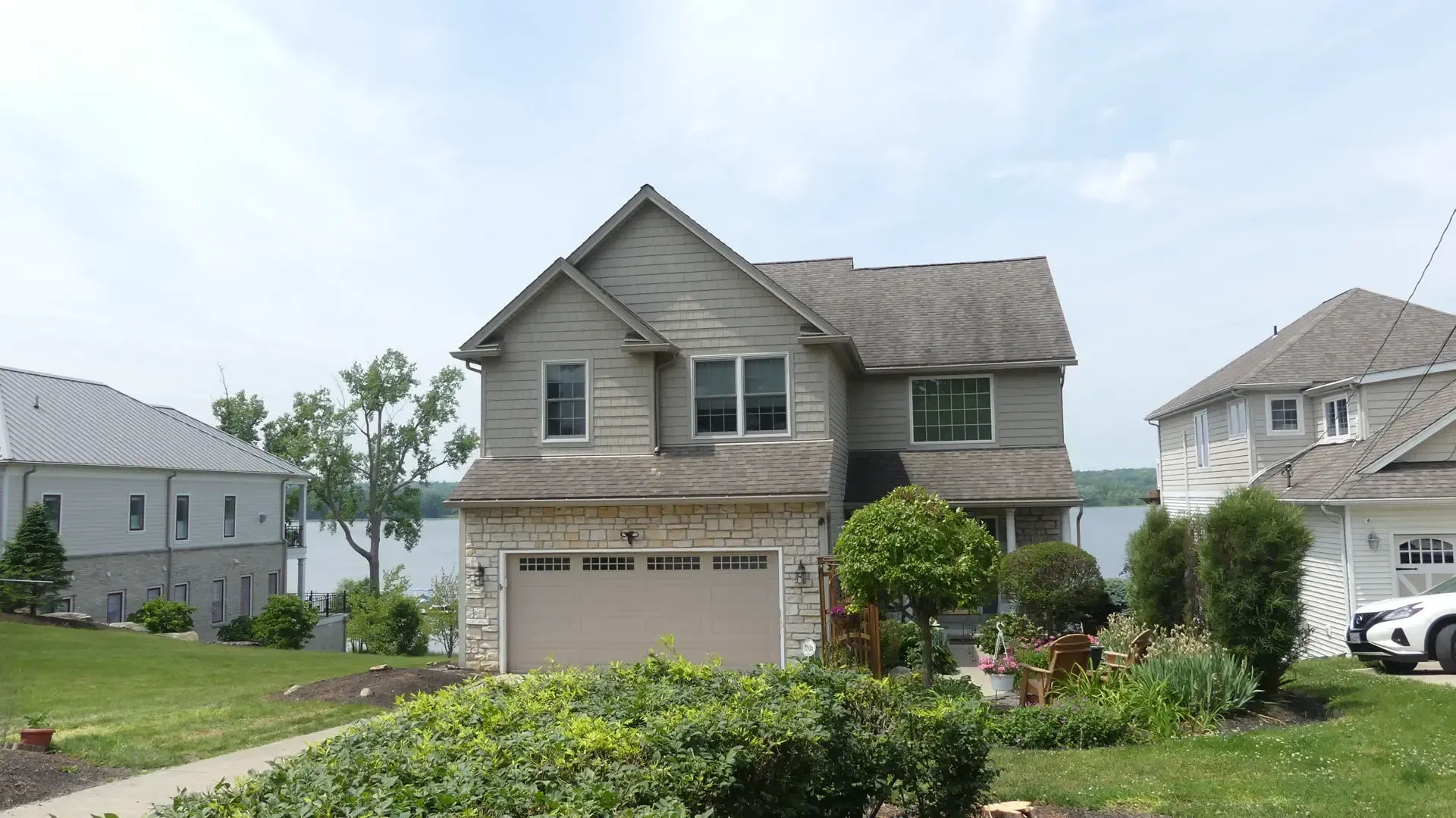A suburban house with beige stone and grey siding, featuring a two-car garage and a lake in the background.