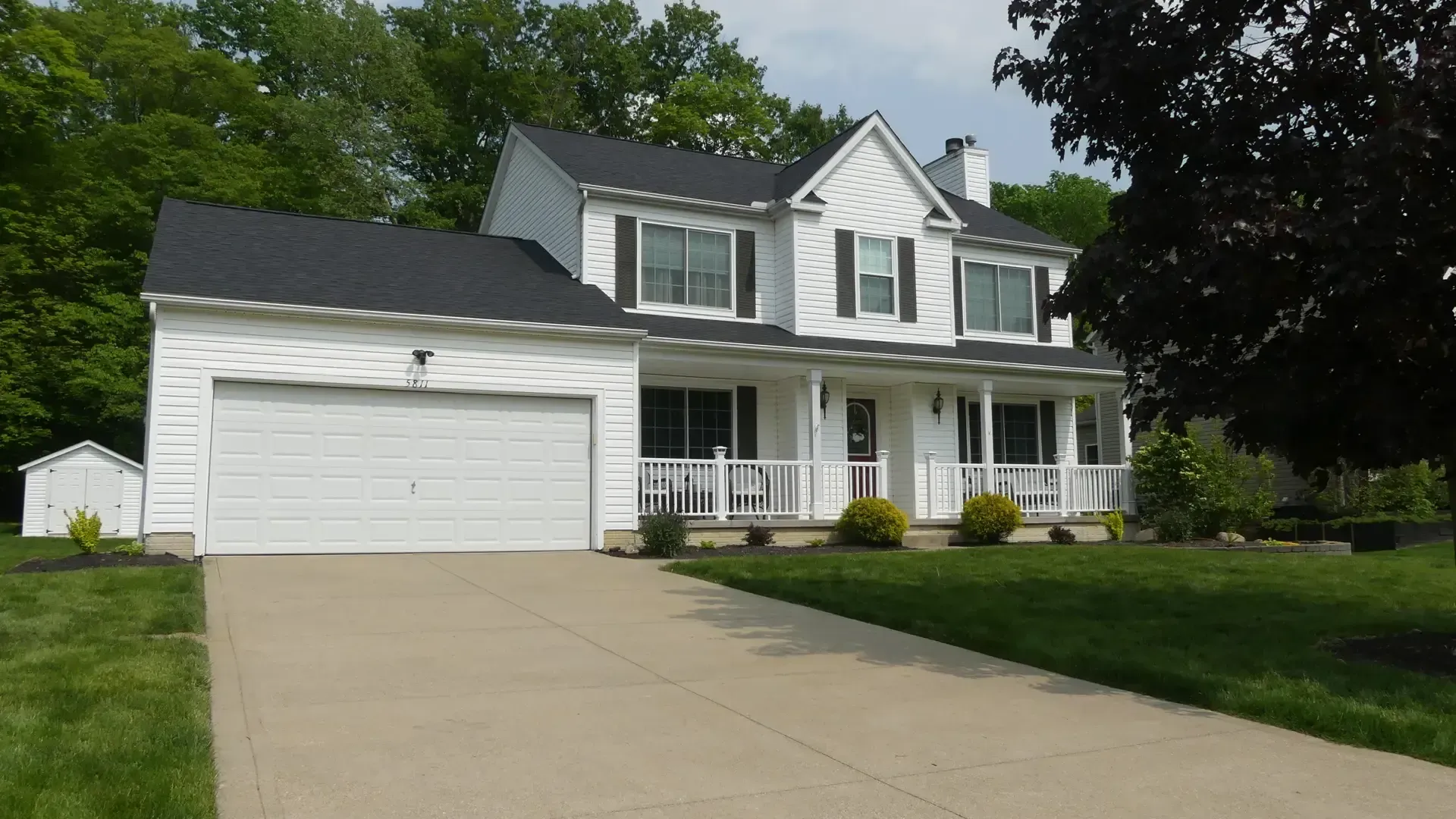 A two-story white house with a black roof, attached garage, and front porch, set on a grassy lot with a concrete driveway.