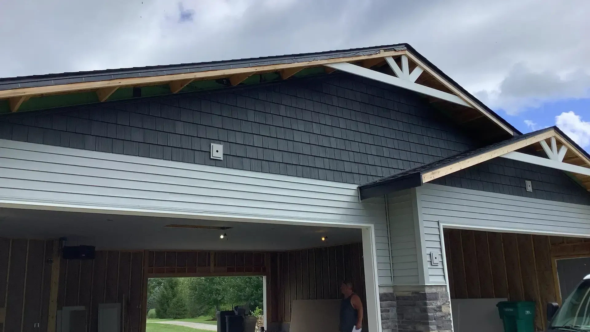 A partially constructed garage with dark shake siding, light horizontal siding, and stone accents under a cloudy sky.