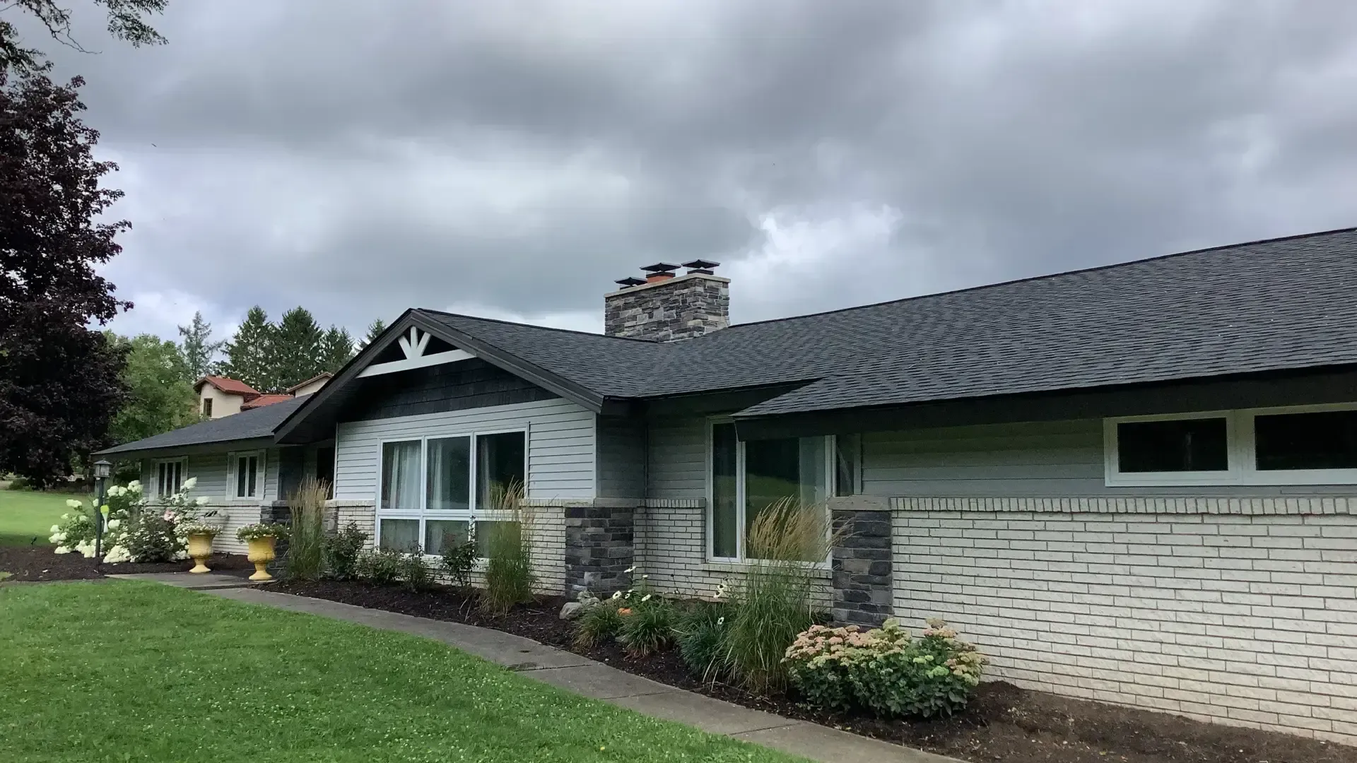 A single-story ranch house with a gray shingled roof, light gray siding, white brick base, and stone chimney on a lawn.