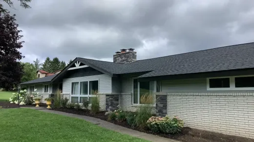 A single-story ranch house with a gray brick exterior, dark shingled roof, stone accents, and a lush front garden.