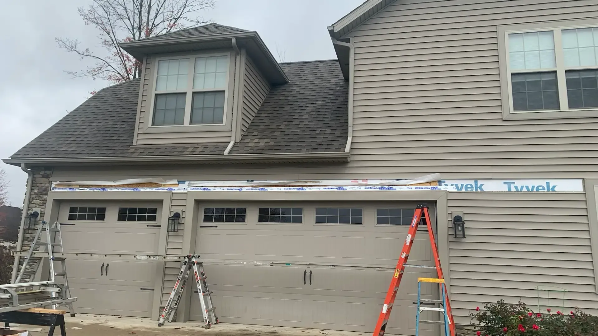 A tan two-story house with a two-car garage under renovation, showing exposed framing and ladders against the exterior.