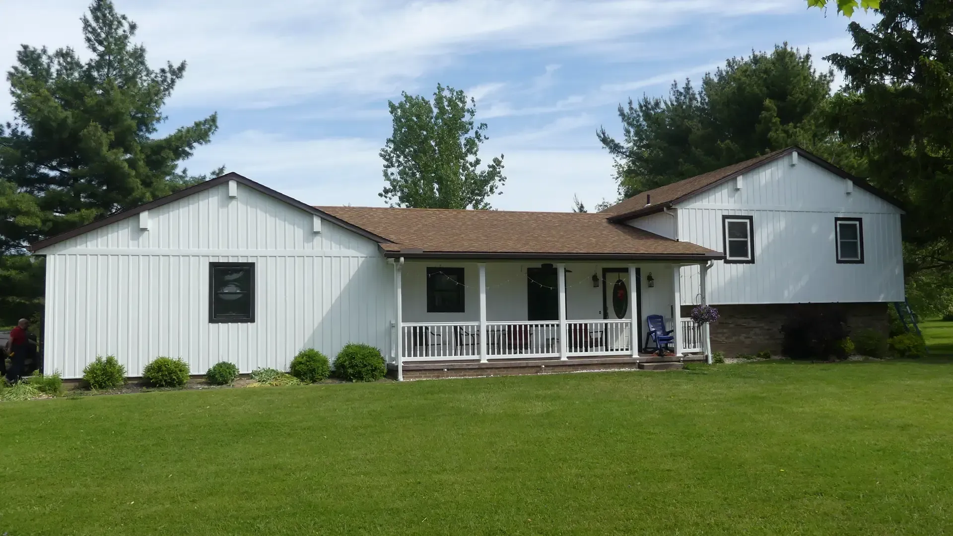A white, two-story farmhouse with vertical siding and a front porch, set against a green lawn and trees.