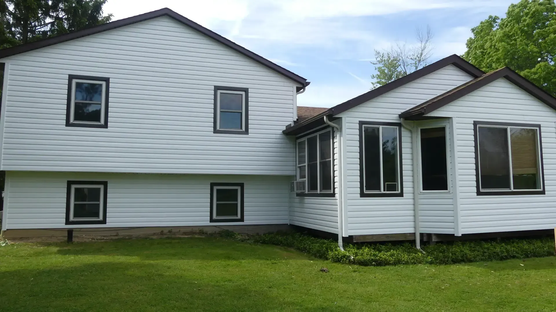 White two-story house with brown trim and horizontal siding, set against a green lawn and trees under a blue sky.