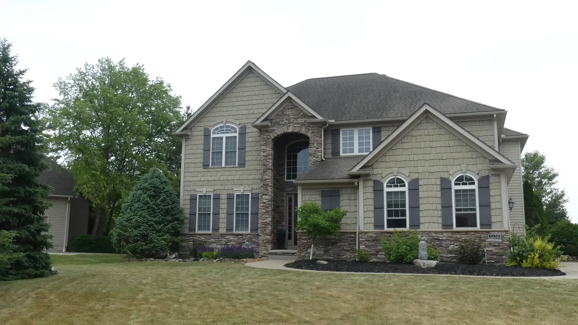 A two-story house with stone and brick exterior, dark shutters, and arched windows, set on a grass lawn with trees.