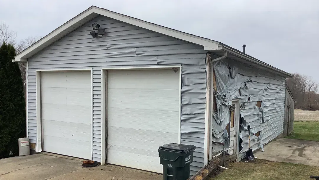 A detached garage with significant exterior siding damage and melting on a cloudy day.