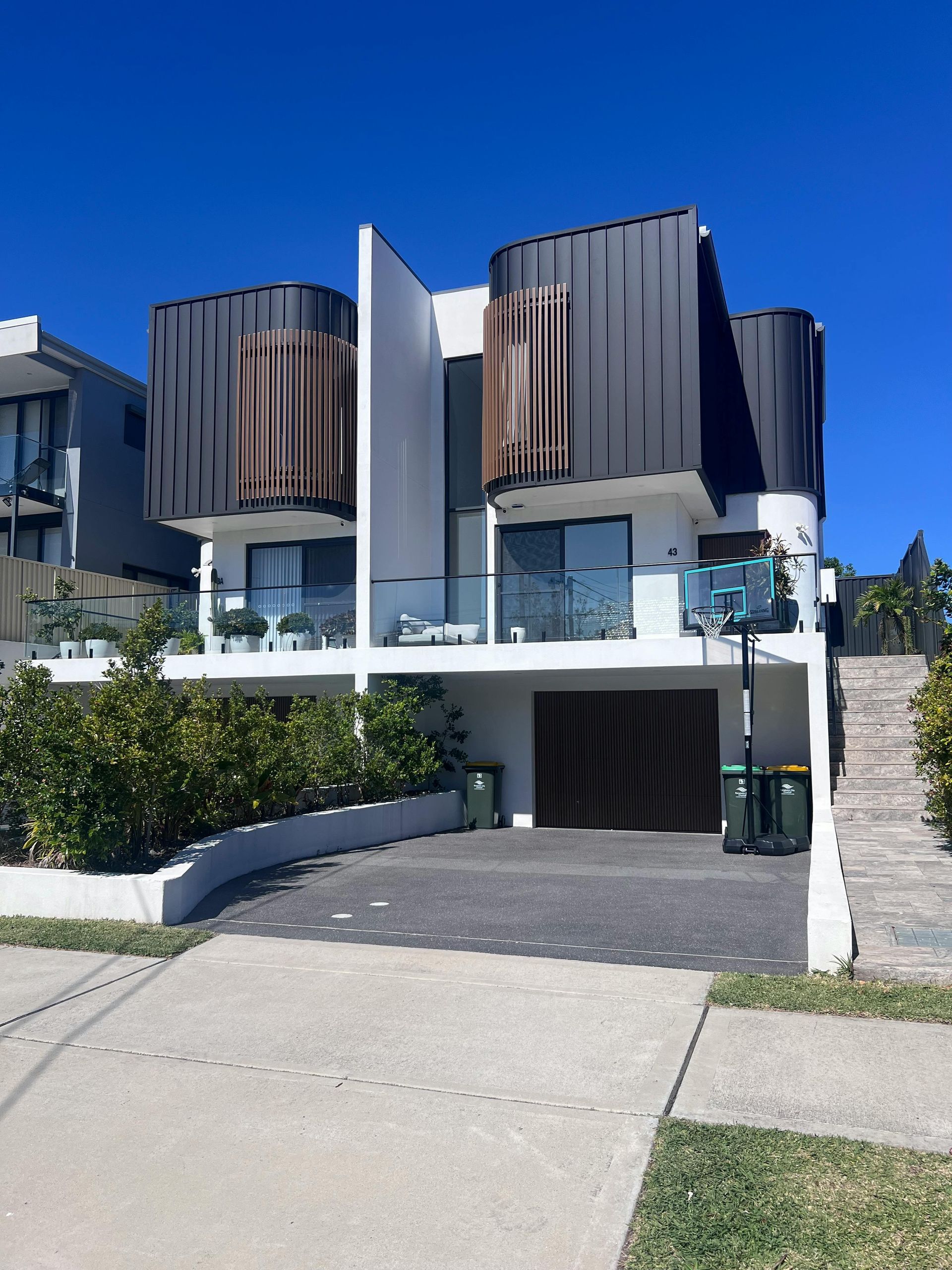 Modern white house with curved dark wood balconies and a driveway under a clear blue sky