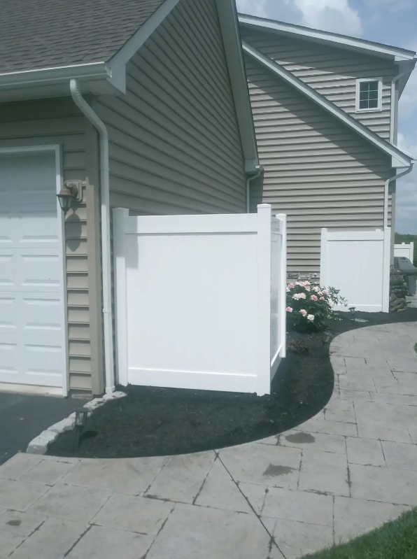 White vinyl fence panels beside a beige house and paved driveway, with a curved walkway and shrubs.