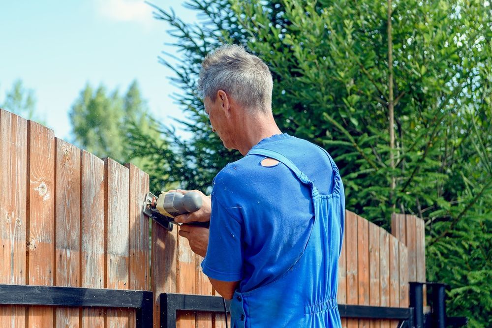 Person in a blue shirt working on a wooden fence outdoors with a power tool